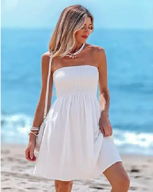 A woman wearing a white strapless tube sundress with a shirred bodice and A-line skirt, standing on a beach.