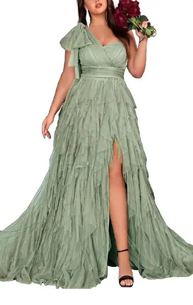 A model wearing a sage green plus-size gown with layered mesh ruffles, an asymmetrical neckline, a decorative shoulder bow, and a high-slit skirt.