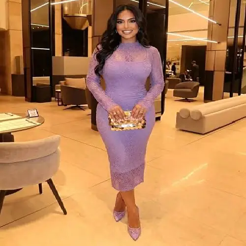 A woman wearing a sophisticated long-sleeve lilac lace midi dress holding a gold clutch.