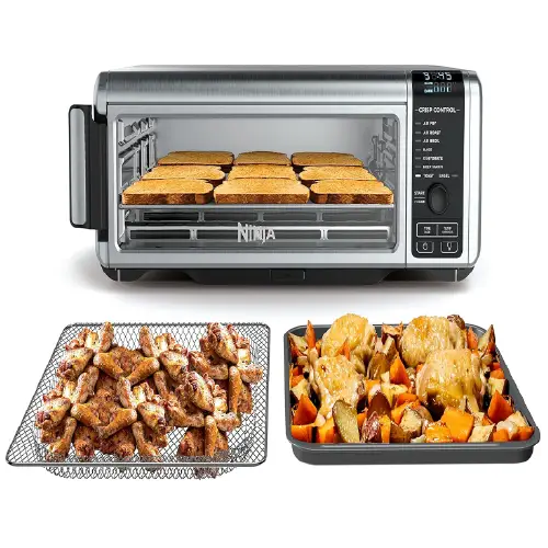 Stainless steel Ninja countertop oven with a long horizontal glass viewing window showing rows of toasted bread, accompanied by a wire air fry basket filled with chicken wings and a tray of roasted vegetables.